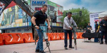 Two men operating jackhammers on a paved plaza at an outdoor event, with orange barriers and sponsor banners in the background, including TAMEX signage.