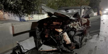 Car crash into concrete barrier at night; front end crushed and debris scattered on the road.