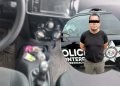 Suspect standing in front of a police vehicle with a star badge visible on the side; eyes covered with a black bar and wearing a black t‑shirt.