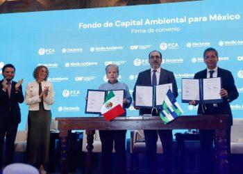 Four officials stand behind a wooden table signing an environmental agreement, Mexican and regional flags visible, blue backdrop reads 'Fondo de Capital Ambiental para México'.
