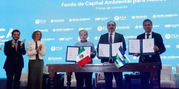 Four officials stand behind a wooden table signing an environmental agreement, Mexican and regional flags visible, blue backdrop reads 'Fondo de Capital Ambiental para México'.