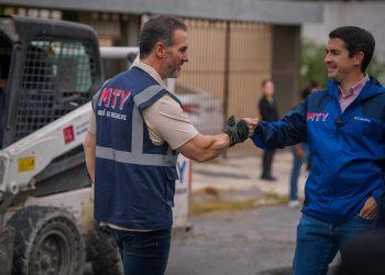 Two workers wearing MTY vests shake hands on a construction site with heavy equipment in the background.