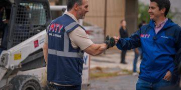 Two workers wearing MTY vests shake hands on a construction site with heavy equipment in the background.