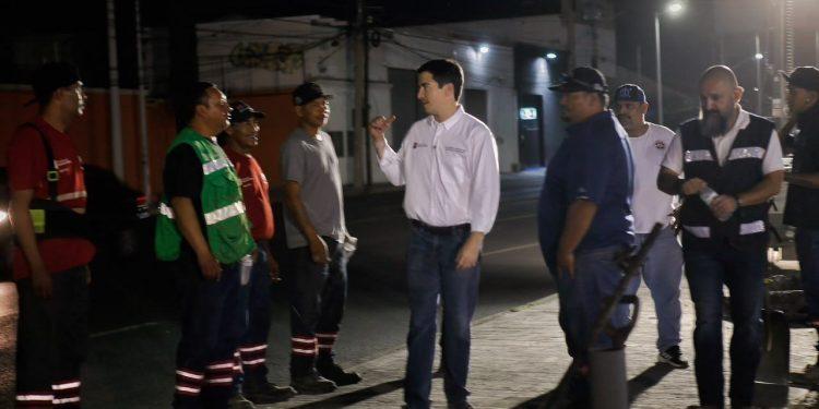 Nighttime street briefing: a man in a white shirt talks to a line of utility workers in reflective vests.